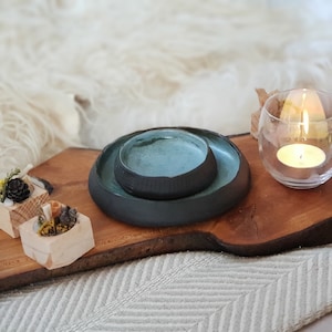 Può includere: Due ciotole in ceramica fatte a mano, una dentro l'altra. La ciotola più grande è grigio scuro, quella più piccola ha un interno turchese. Sono su una tavola di legno, con una candela accesa in un portacandele di vetro.