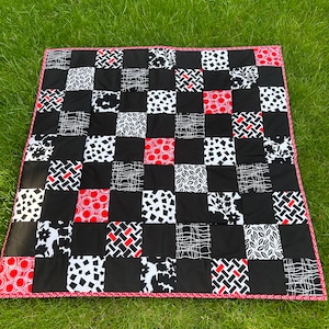 May include: A handmade quilt with a patchwork design. The quilt features a black and white checkered pattern with red accents. The border is a pink and white dotted fabric. The quilt is displayed on a green grassy surface.