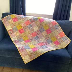 May include: A patchwork quilt with a yellow trim is draped over a blue sofa. The quilt is made up of many fabric squares in shades of pink, yellow, and lilac. The squares have floral and patterned designs.