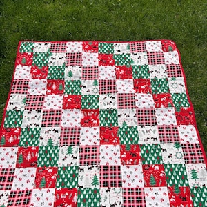 May include: A red, green and white patchwork quilt with a Christmas theme. The quilt is made up of squares with different patterns, including plaid, snowflakes and Christmas trees. The quilt is lying on a green grassy surface.