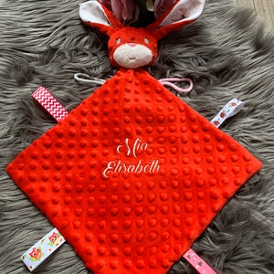 Könnte beinhalten: Rote Baby-Sicherheitsdecke mit einem daran befestigten Hasenkopf. Die Decke hat eine strukturierte Oberfläche und ist mit dem Namen "Mia Elizabeth" bestickt.