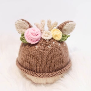 May include: A brown knit baby hat with deer ears and small antlers. The hat is decorated with pink, yellow, and white flowers and green leaves. The hat is designed to look like a deer.