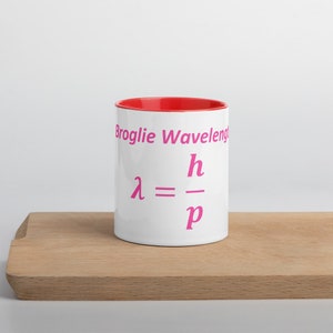 May include: A white ceramic mug with a bright red interior. The exterior features the pink text Broglie Wavelength and the physics equation lambda equals h divided by p printed in a matching pink font on the front of the cylindrical vessel.