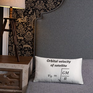 May include: A rectangular white throw pillow featuring the black printed text Orbital velocity of satellite and the physics formula v0 equals the square root of GM divided by R. The pillow has a smooth fabric surface and a clean, simple design.