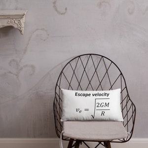 May include: A rectangular white throw pillow featuring the black printed text Escape velocity and the physics formula v_e = square root of 2GM/R. The pillow is positioned on a dark metal wire chair with a light-colored seat cushion.