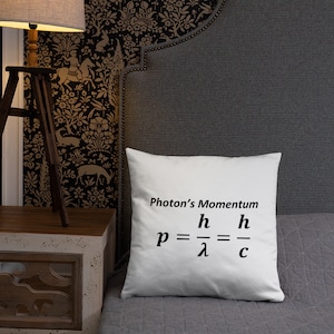 Op de afbeelding: Een vierkant wit sierkussen met zwarte bedrukte tekst die Photon's Momentum leest, gevolgd door de natuurkundige vergelijking p = h/λ = h/c. Het kussen heeft een glad stoffen oppervlak en staat tegen een donkergrijs gestoffeerd hoofdeinde.