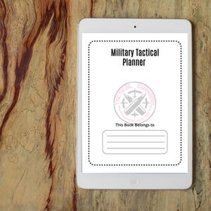 May include: A white tablet displays a "Military Tactical Planner" cover. The cover includes a logo with crossed bullets and the text "This Book Belongs to" with lines for the owner's name. The tablet rests on a wooden surface.