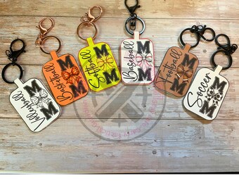 May include: Keychains with sports themes, including volleyball, basketball, softball, baseball, football, and soccer. Each keychain features a rectangular design with the word "MOM" and a sports-related graphic. The keychains have different colored backgrounds and metal hardware.