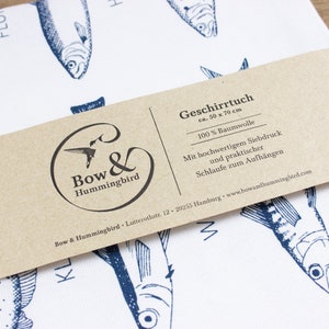 May include: A brown paper tag with white text that reads "Geschirrtuch ca. 50 x 70 cm 100% Baumwolle Mit hochwertigem Siebdruck und praktischer Schlaufe zum Aufh&auml;ngen Bow & Hummingbird Lutterothstr. 12. 20255 Hamburg www.bowandhummingbird.com"  The tag is attached to a white tea towel with a blue and white fish pattern.