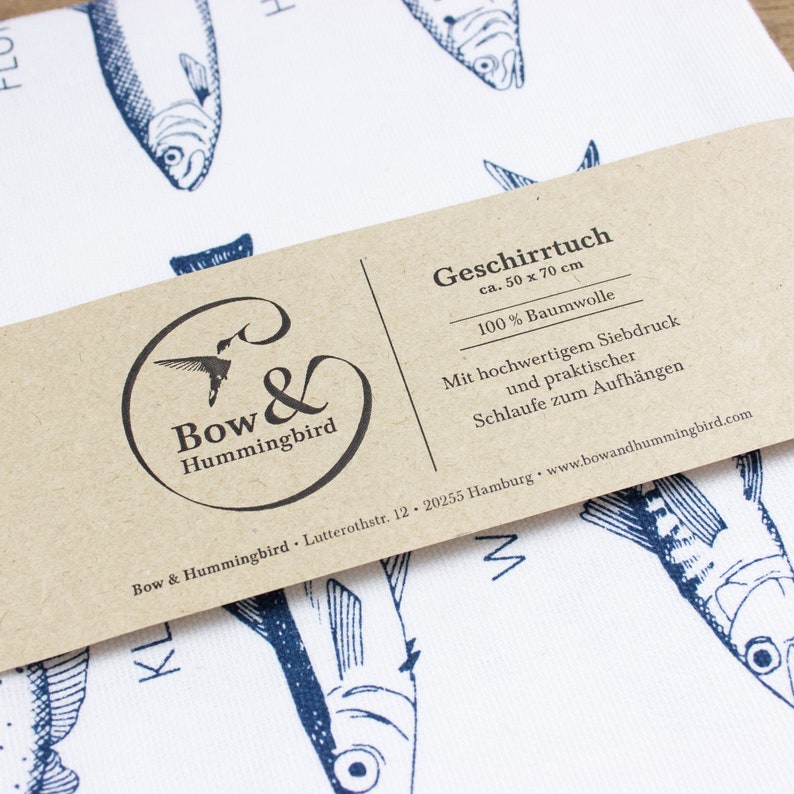 May include: A brown paper tag with white text that reads "Geschirrtuch ca. 50 x 70 cm 100% Baumwolle Mit hochwertigem Siebdruck und praktischer Schlaufe zum Aufh&auml;ngen Bow & Hummingbird Lutterothstr. 12. 20255 Hamburg www.bowandhummingbird.com"  The tag is attached to a white tea towel with a blue and white fish pattern.
