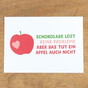 Könnte beinhalten: Ein roter Apfel mit einem rosa Herzen auf weißem Hintergrund. Der Text "SCHOKOLADE LÖST KEINE PROBLEME ABER DAS TUT EIN APFEL AUCH NICHT" ist in grün und rosa geschrieben.