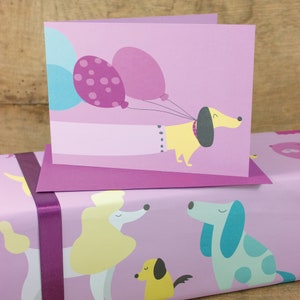 May include: A purple card with a yellow dachshund holding three purple balloons. The card is on top of a purple wrapping paper with a pattern of dogs.