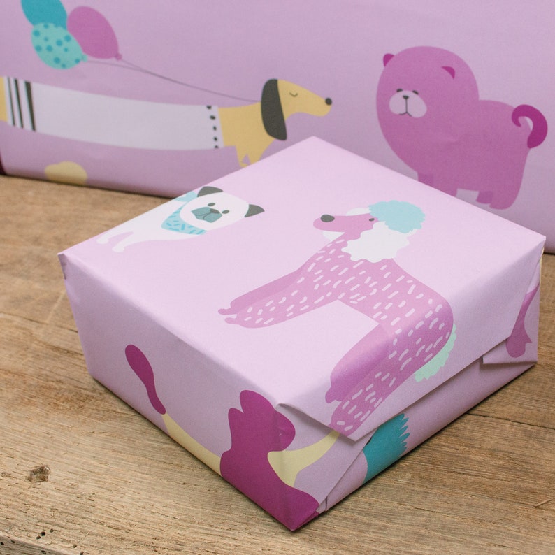 May include: A pink gift box with a wrapping paper design featuring various cartoon dogs. The dogs are a pug, a poodle, and a dachshund. The box is wrapped in a light pink paper with a white border.