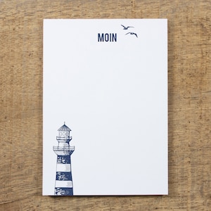 Könnte beinhalten: Ein weißer Notizblock mit einer blauen Leuchtturm-Illustration und dem Wort "MOIN" obenauf gedruckt. Zwei blaue Vögel fliegen über dem Leuchtturm.