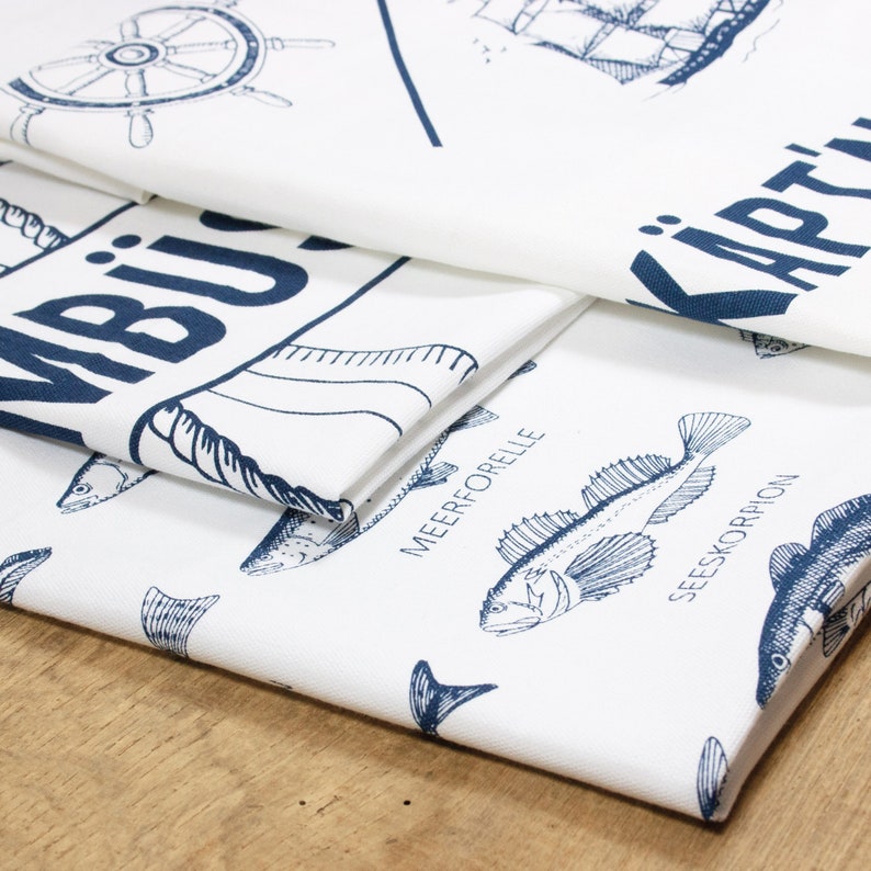 May include: Three white tea towels with blue nautical designs. The top towel features a ship's wheel and the text "KARTEN". The middle towel has the text "MBIJ" and a stylized fish design. The bottom towel has a pattern of various fish with their names in German: "MEERFORELLE", "SEESKORPION", and "SKORPION".