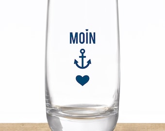 Pode incluir: Um copo transparente com base arredondada. O copo apresenta a palavra "MOIN" em azul marinho, juntamente com uma âncora e um coração. O copo está sobre uma superfície de madeira.