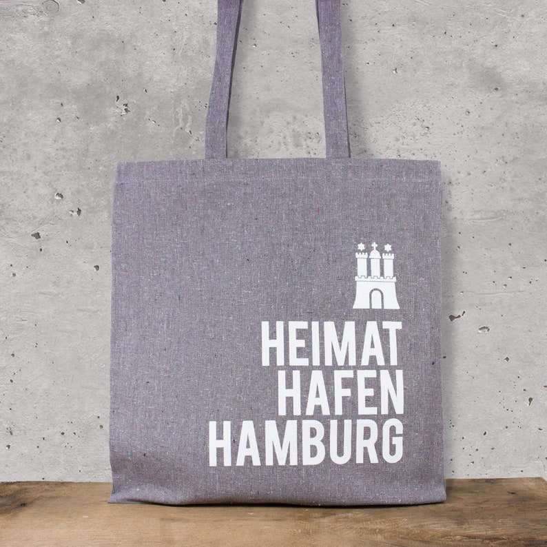 May include: A gray tote bag with white text that reads "Heimat Hafen Hamburg" and a white silhouette of a castle with three towers.