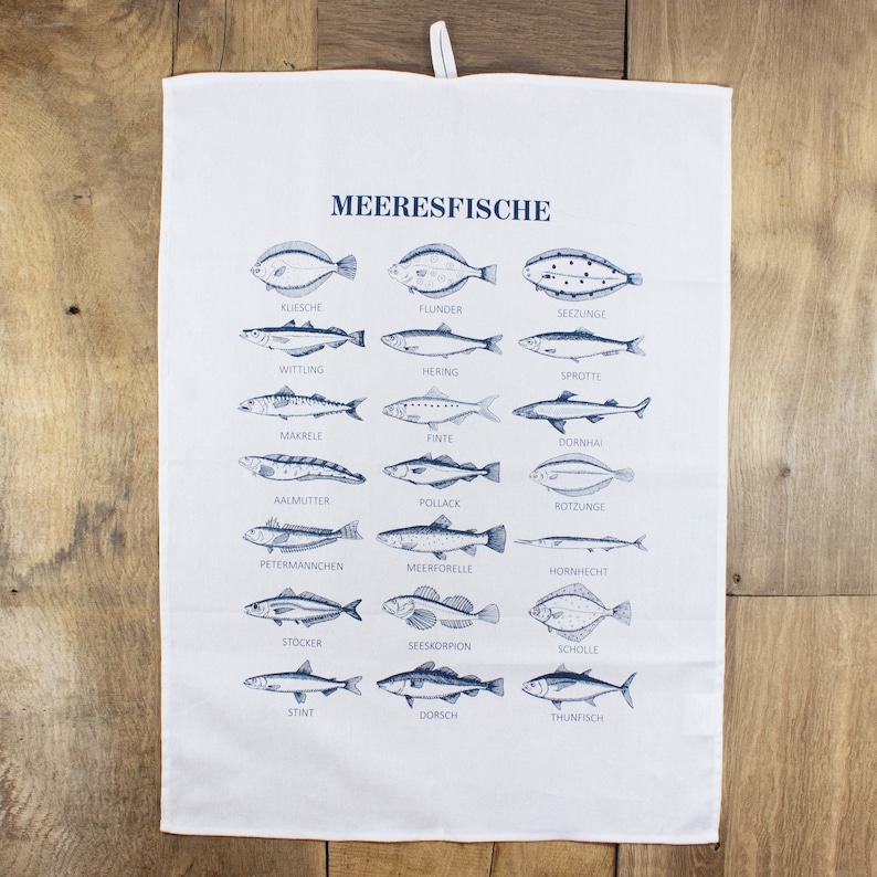 May include: A white kitchen towel with a blue illustration of different types of fish. The names of the fish are written in German below each illustration. The text at the top of the towel reads "MEERESFISCHE".