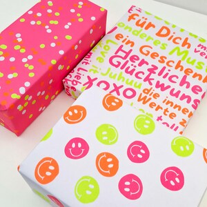 Pode incluir: Tr&ecirc;s caixas de presente embrulhadas. Uma &eacute; rosa brilhante com confetes laranja neon e branco. Outra tem fundo branco com carinhas sorridentes em laranja neon, rosa e verde. A terceira caixa tem texto em laranja neon e rosa.