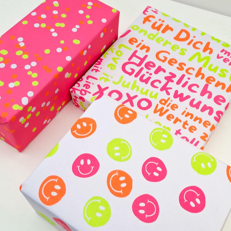 Pode incluir: Tr&ecirc;s caixas de presente embrulhadas. Uma &eacute; rosa brilhante com confetes laranja neon e branco. Outra tem fundo branco com carinhas sorridentes em laranja neon, rosa e verde. A terceira caixa tem texto em laranja neon e rosa.