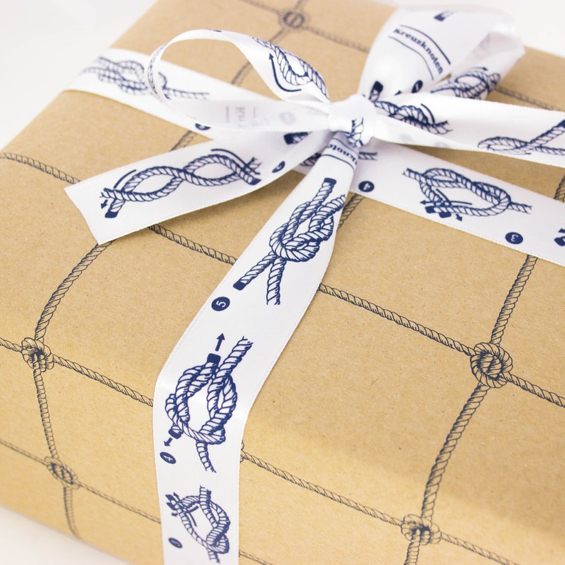 Puede incluir: Una caja de regalo de papel marr&oacute;n con una cinta blanca atada en un lazo. La cinta tiene un patr&oacute;n de nudos marinos azules con n&uacute;meros y flechas. La caja est&aacute; envuelta en papel marr&oacute;n con un patr&oacute;n de cuerda azul.