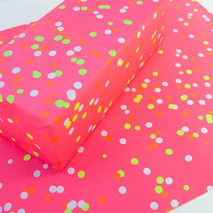 Pode incluir: Papel de embrulho rosa brilhante com um padr&atilde;o de confetes em branco, laranja e verde n&eacute;on. Um presente retangular embrulhado est&aacute; sobre uma folha do mesmo papel, criando uma apresenta&ccedil;&atilde;o festiva e colorida.