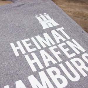 May include: Gray tote bag with white text that reads "Heimat Hafen Hamburg" and a white silhouette of a castle.