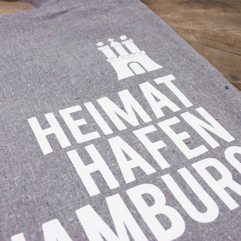 May include: Gray tote bag with white text that reads "Heimat Hafen Hamburg" and a white silhouette of a castle.