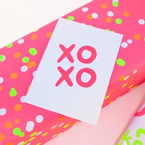 Pode incluir: Um cart&atilde;o branco com o texto "XO XO" em rosa brilhante, colocado em cima de um papel de embrulho rosa com bolinhas coloridas. O papel de embrulho &eacute; rosa vibrante com pontos laranja, verdes e brancos.