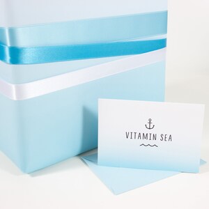Könnte beinhalten: Eine hellblaue Geschenkbox mit weißem und blauem Band und einer weißen Karte mit dem Text "VITAMIN SEA" und einem Anker-Symbol.