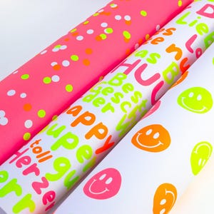 Pode incluir: Tr&ecirc;s rolos de papel de embrulho colorido. Um &eacute; rosa com confetes, outro &eacute; branco com texto laranja n&eacute;on, rosa e verde, e o terceiro &eacute; branco com carinhas sorridentes e detalhes em verde n&eacute;on.