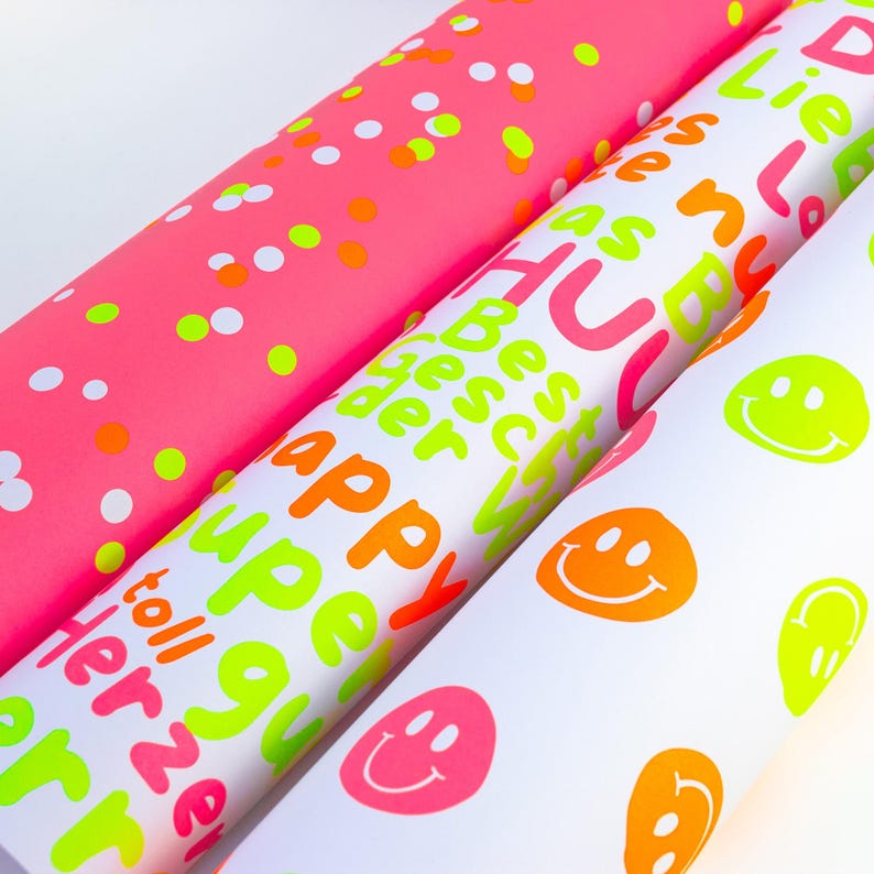 Pode incluir: Tr&ecirc;s rolos de papel de embrulho colorido. Um &eacute; rosa com confetes, outro &eacute; branco com texto laranja n&eacute;on, rosa e verde, e o terceiro &eacute; branco com carinhas sorridentes e detalhes em verde n&eacute;on.