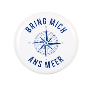 Könnte beinhalten: Weißer runder Knopf mit blauem Text und einem Kompass-Design. Der Text lautet "Bring mich ans Meer".