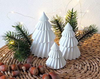 May include: Three white ceramic Christmas trees of varying sizes, with the largest tree in the back and the smallest tree in the front. The trees are sitting on a woven brown basket with nuts scattered around them.