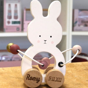 Könnte beinhalten: Weißes Holz-Ziehspielzeug in Form eines Hasen mit rosa und goldenen Perlen an einer Schnur. Der Hase hat schwarze Augen und eine schwarze Nase. Die Räder sind mit dem Namen "Romy" und dem Datum "17.02.2022" graviert.