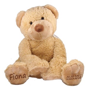 Könnte beinhalten: Ein beiger Plüschteddybär mit braunen Augen und einer braunen Nase. Der Bär sitzt mit seinen Pfoten vor sich. Die linke Pfote des Bären hat den Namen "Fiona" gestickt und die rechte Pfote das Datum "01.01.2016" gestickt.