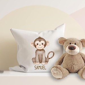 Könnte beinhalten: Ein weißes quadratisches Kissen mit einer Aquarellillustration eines Affen und dem Namen "SAMUEL" darunter. Ein brauner Teddybär befindet sich neben dem Kissen. Die Affenillustration ist in Braun- und Beigetönen gehalten.