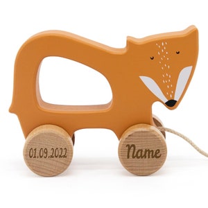 Könnte beinhalten: Orangefarbenes Holz-Ziehspielzeug in Form eines Fuchses mit weißem Gesicht und schwarzer Nase. Das Spielzeug hat zwei Holzräder mit dem eingravierten Text "01.09.2022" und "Name".
