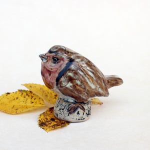 Op de afbeelding: Een keramieken vogelbeeldje met bruin-witte gestreepte veren, een roze borst en een donkerblauwe streep over het oog. De vogel zit op een getextureerde basis met gele bladeren.