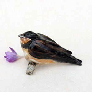 Op de afbeelding: Een keramieken vogelbeeldje met een zwarte kop en vleugels, een bruine borst en een bruine rug. De vogel zit op een kleine tak naast een paarse bloem. Het beeldje staat op een witte achtergrond.