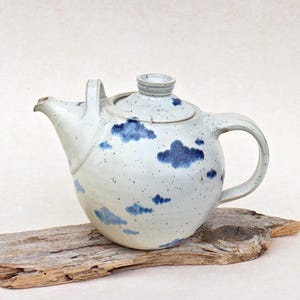 Può includere: Una teiera rotonda in ceramica con smalto bianco sporco e finitura maculata. La teiera presenta un motivo dipinto a mano di piccole nuvole blu scuro sparse sul corpo e sul coperchio. È dotata di un manico curvo e di un beccuccio corto e affusolato.