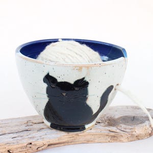 Può includere: Ciotola per filato in ceramica fatta a mano con un disegno di gatto nero. La ciotola è bianca con dettagli blu e un interno blu. Una palla di filo bianco è all'interno, con il filo che esce da un foro laterale. La ciotola è appoggiata su un pezzo di legno.