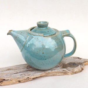 Può includere: Una teiera in ceramica turchese con corpo arrotondato, beccuccio e manico. La teiera ha un coperchio e poggia su un pezzo di legno alla deriva. La ceramica ha un aspetto maculato.