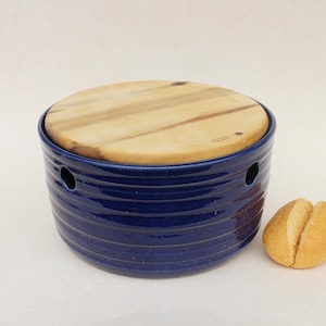 Könnte beinhalten: Eine blaue Keramik-Butterdose mit Holzdeckel. Die Dose hat eine runde Form mit horizontalen Rillen und zwei kleinen Löchern. Eine kleine Brötchen liegt neben der Dose.