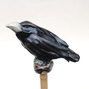 Op de afbeelding: Een zwarte keramische ravenfiguur met een glanzende afwerking en een lichtgrijze snavel. De raaf zit op een kleine, getextureerde stenen basis die aan een houten deuvel is bevestigd. De vogel is iets omhoog gekanteld.