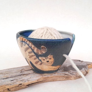 Può includere: Una ciotola per filato in ceramica blu con un disegno di gatto. La ciotola contiene una palla di filo bianco e un filo esce attraverso una fessura. La ciotola è appoggiata su un pezzo di legno.
