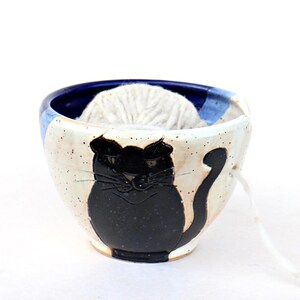 Pode incluir: Uma tigela de cerâmica para fios com um desenho de gato preto. A tigela é branca com detalhes azuis e uma silhueta de gato preto. Uma bola de fio branco está dentro, com um fio saindo. Ideal para tricô e crochê.