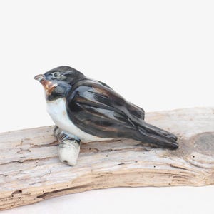 Op de afbeelding: Een keramieken vogelbeeldje met een glanzende afwerking, zittend op een stuk drijfhout. De vogel heeft zwarte, bruine, witte en oranje kleuren. Het beeldje is een decoratief item, geschikt voor woondecoratie.