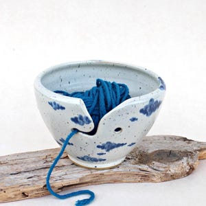 Può includere: Una ciotola per lana in ceramica maculata con base grigio chiaro e nuvole blu scuro dipinte a mano. La ciotola presenta un taglio a spirale per il filo e un piccolo foro circolare sul lato, contenente un gomitolo di lana blu intenso.