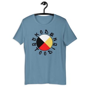 May include: A light blue t-shirt with a black circle design. The circle contains a four-color symbol with black, white, red, and yellow sections. The text "TODAS KODEN DISN" is written around the circle.